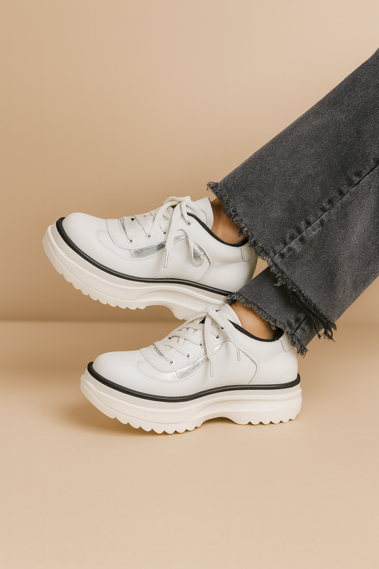 Zapatilla de cuero blanca con detalle plateado en el lateral, mujer con jeans grises sobre fondo beige, suela plataforma dentada.