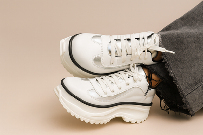 Primer plano de zapatilla de cuero blanca con detalle plateado y cordones blancos, jeans grises deshilachados, suela dentada.