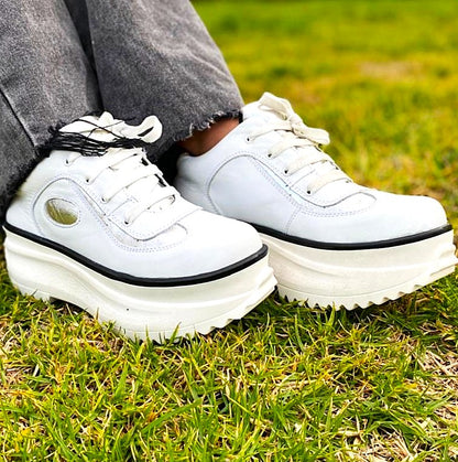 Par de zapatillas de cuero blancas con detalle plateado, usadas por mujer sobre pasto verde en exterior, plataforma gruesa dentada.